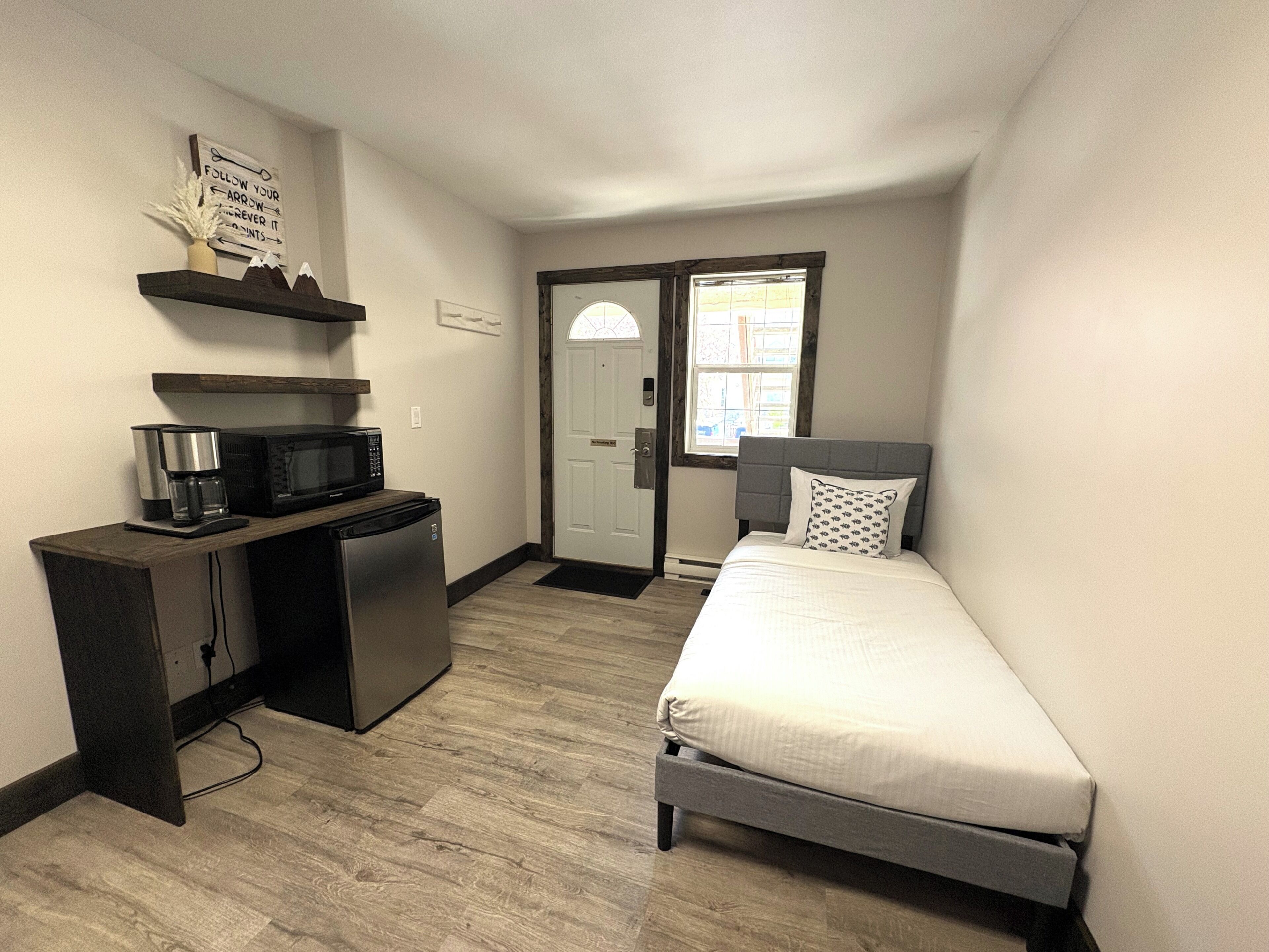 A compact room with a bed, a desk with a microwave, a refrigerator, and a door leading to a balcony.