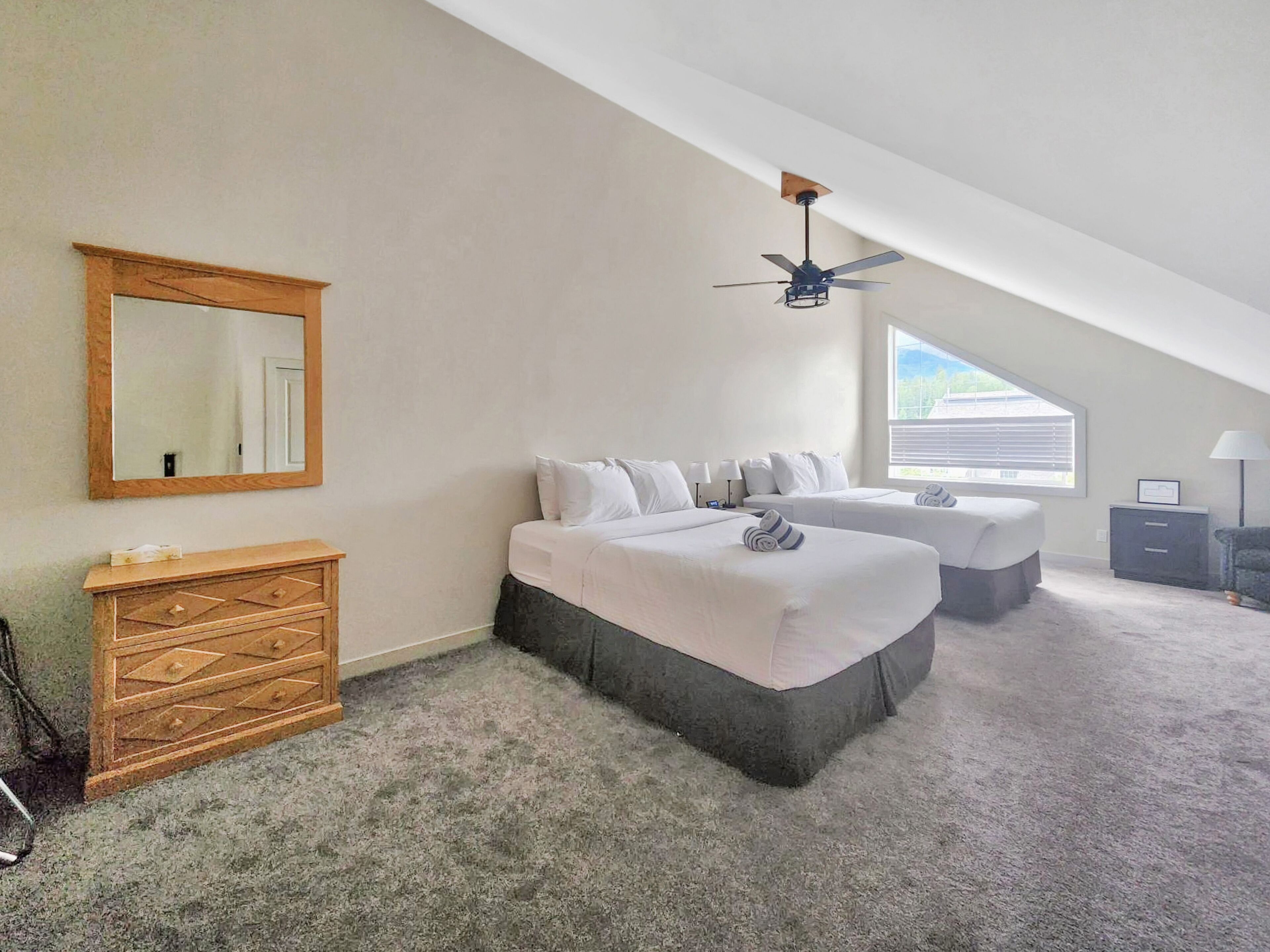 A bedroom with two beds, a ceiling fan, a large window, and a dresser.