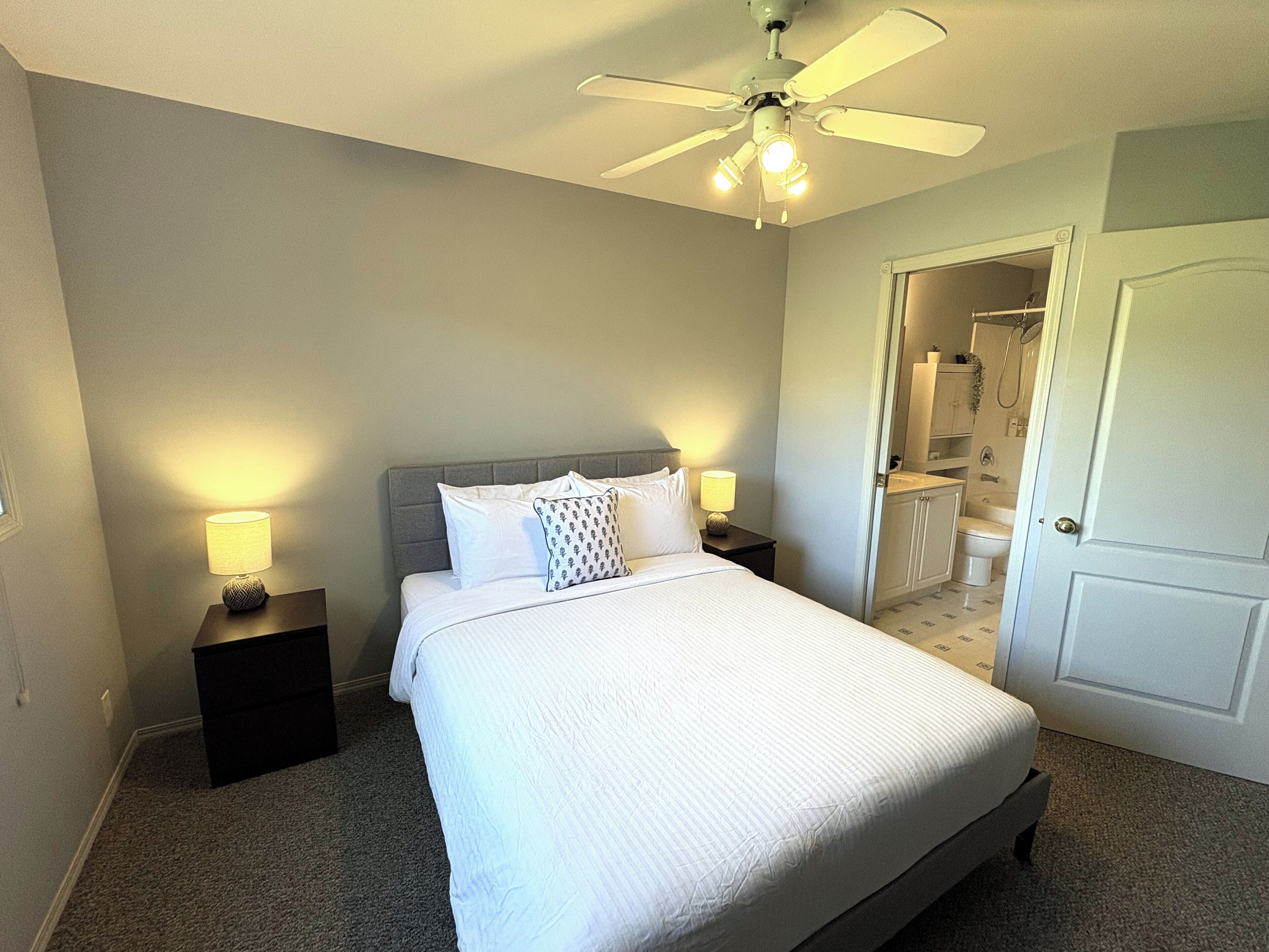 A neatly made bed with a headboard, two bedside lamps, a ceiling fan, and a bathroom visible through an open door.