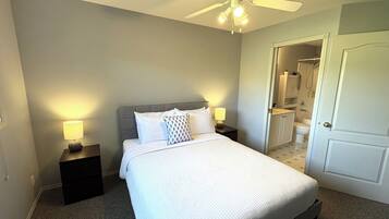 A neatly made bed with a headboard, two bedside lamps, a ceiling fan, and a bathroom visible through an open door.