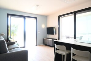 Una sala de estar moderna con un sofá, una mesa de comedor con sillas, un televisor de pantalla plana montado en la pared y ventanas amplias.