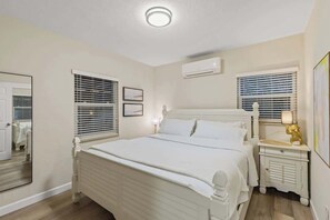 A bedroom with a bed, bedside tables, a mirror, and an air conditioning unit.