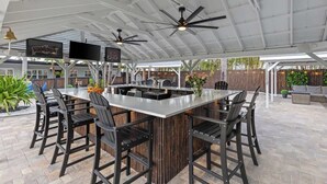 Un área de bar al aire libre con una isla central, varias sillas, ventiladores de techo y una pantalla de televisión.