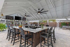 Un área de bar al aire libre con una isla central, varias sillas, ventiladores de techo y una pantalla de televisión.