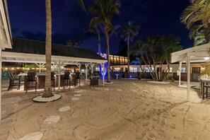 A beachside outdoor seating area with tables and chairs, palm trees, and a building with illuminated windows in the background.
