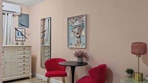 Una habitación con un aparador blanco, dos sillas rojas, una mesa redonda con una planta en maceta, un cuadro en la pared y una lámpara de pie.