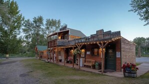 A rustic wooden building with a porch, string lights, and a flower bed. - Sheriff's Office Cabin At Mountain River Ranch (Ririe)