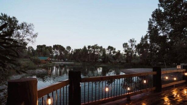 A wooden deck with a railing overlooking a calm body of water, surrounded by trees and a distant building. - Sheriff's Office Cabin At Mountain River Ranch (Ririe)
