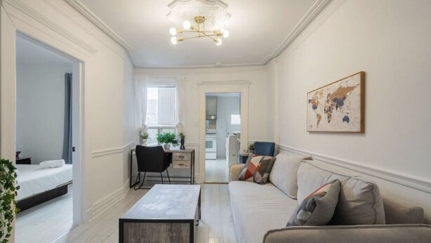 A spacious living room with a sofa, a coffee table, and a desk with a chair. There is a large window providing natural light and a view into the kitchen.