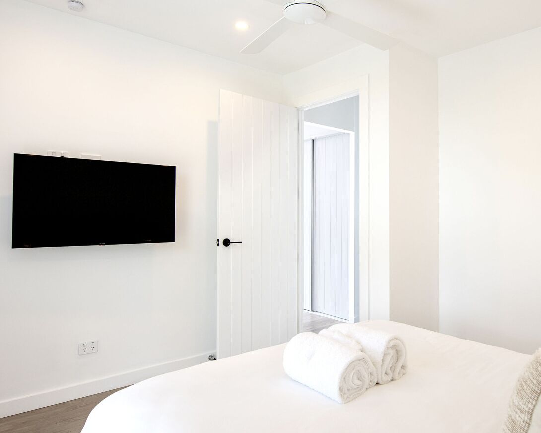 Ein modernes Hotelzimmer mit einem an der Wand befestigten Flachbildfernseher, einer weißen Tür und einem ordentlich bezogenen Bett mit gerollten weißen Handtüchern.