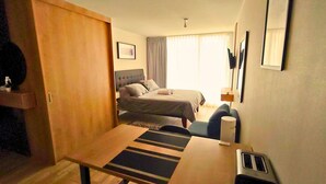 Habitación de hotel compacta con cama, escritorio, silla, mesita, espejo y ventana con cortinas.