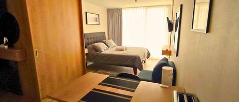 Habitación de hotel compacta con cama, escritorio, silla, mesita, espejo y ventana con cortinas.