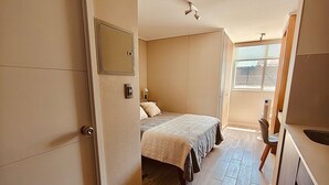 A compact bedroom with a bed, a bedside table, a mirror, and a window. - Confort de Ñuñoa (Santiago)