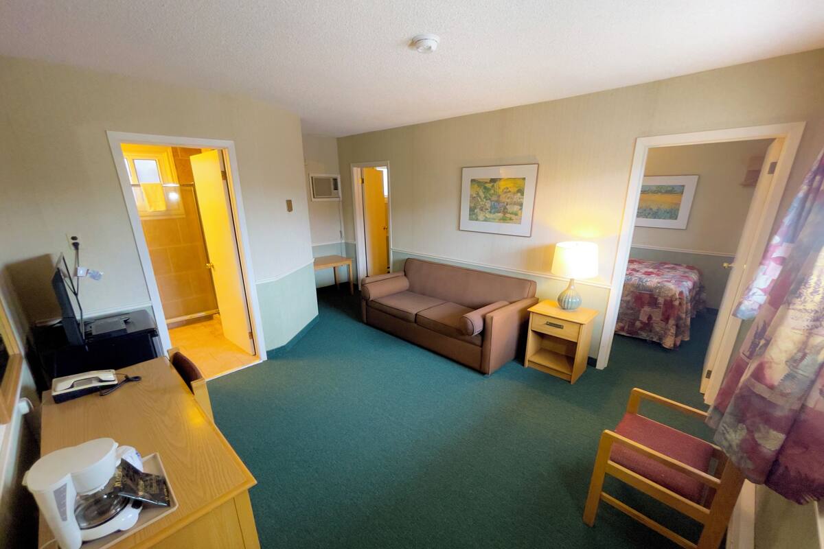 A compact hotel room with a sofa, a small table with a lamp, a chair, and a bed with a quilt.