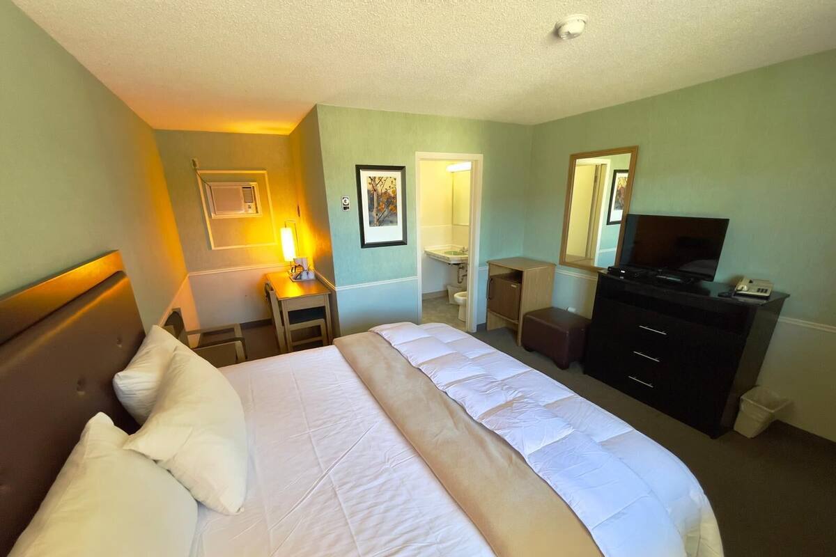A hotel room with a large bed, a desk with a lamp, a dresser, a television, and a bathroom visible through an open door.