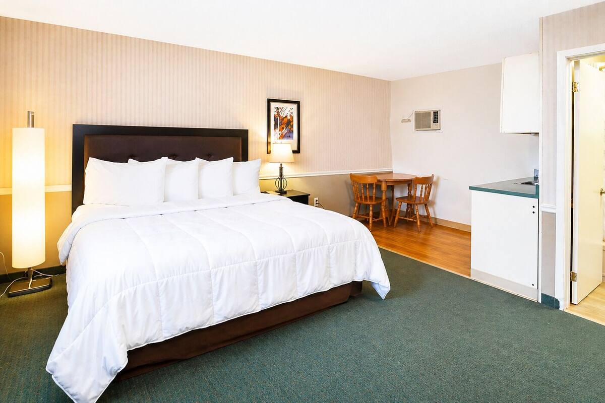A hotel room with a bed, a desk, a chair, a small table, a lamp, and a bathroom door.