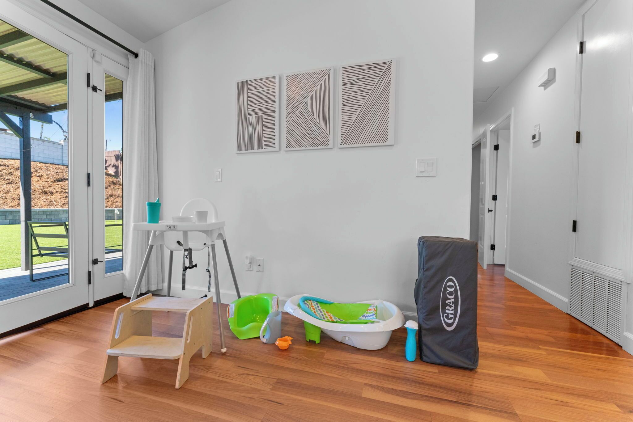 Una habitación con piso de madera, un cambiador para bebés, una bañera para bebés, un contenedor verde de almacenamiento y un gimnasio para bebés GRACO.