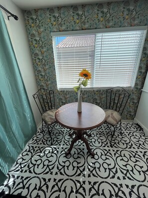 A room with a patterned floor, a round table with a vase of flowers, and a window with blinds.