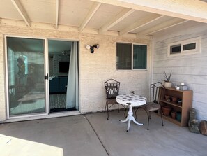 A covered patio area with a sliding glass door leading to a living room, a round table with chairs, and a shelf with potted plants.