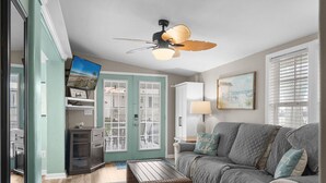 A living room with a ceiling fan, a television, a couch, and a wicker coffee table. - Cottage in Ocean Lakes w/ Gated Porch + Golf Cart (Myrtle Beach)
