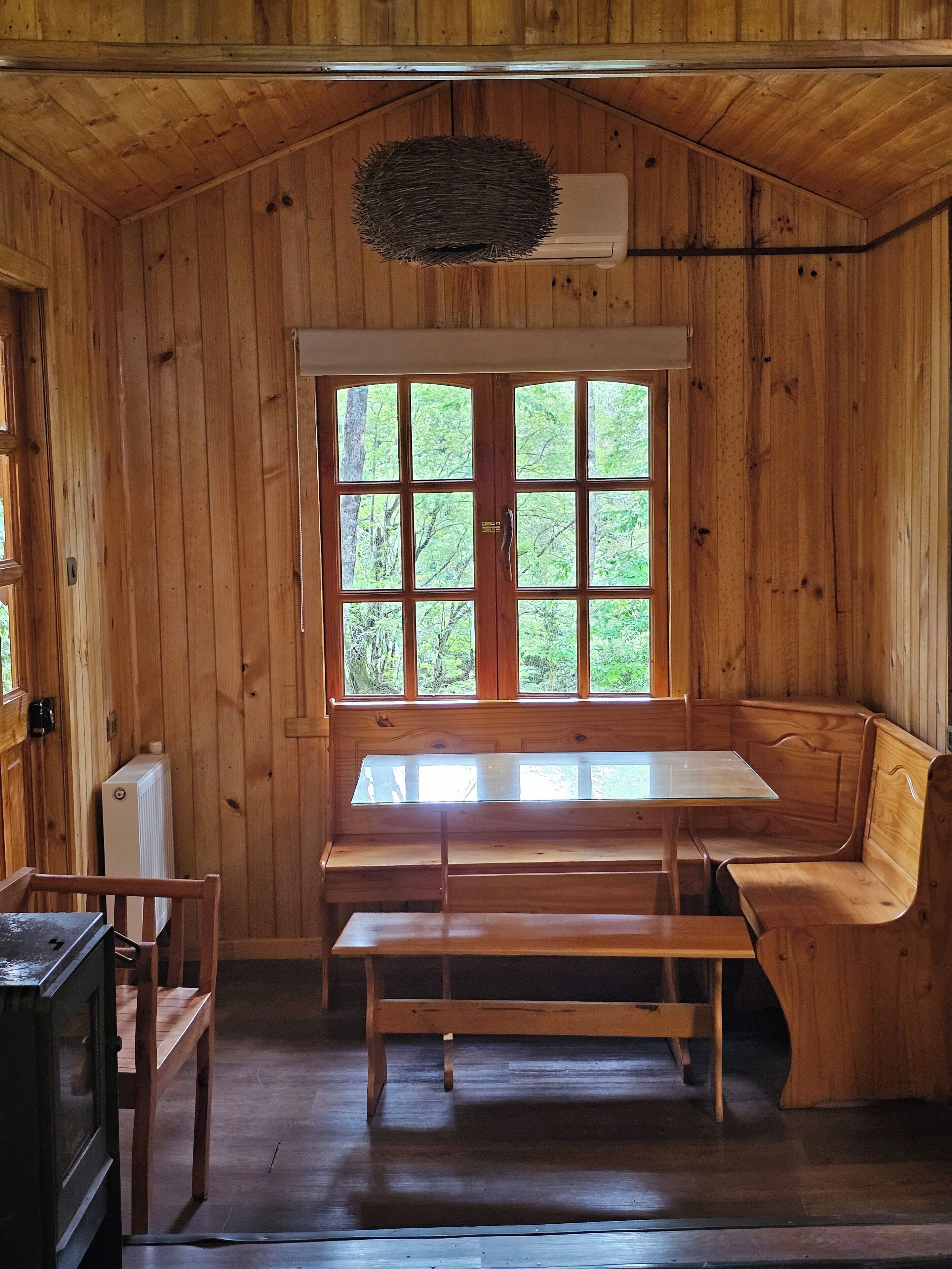 A wooden cabin interior with a large window, a bench, a table, and a radiator.