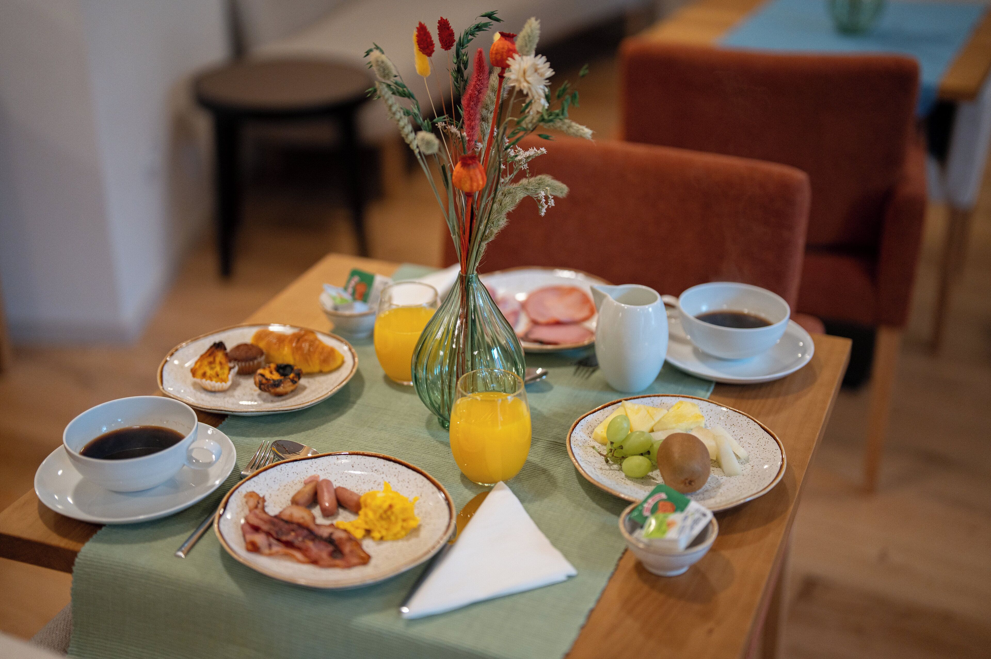A breakfast table with plates of eggs, bacon, toast, and fruit, along with a vase of flowers and a glass of orange juice.