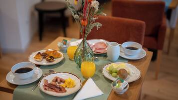 A breakfast table with plates of eggs, bacon, toast, and fruit, along with a vase of flowers and a glass of orange juice.