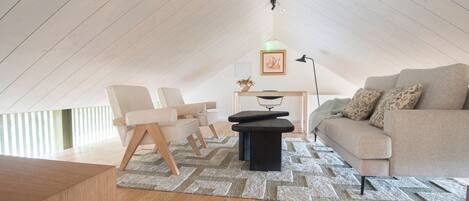 A modern loft bedroom with a sloped ceiling, a sofa, a chair, a small table, and a floor lamp.