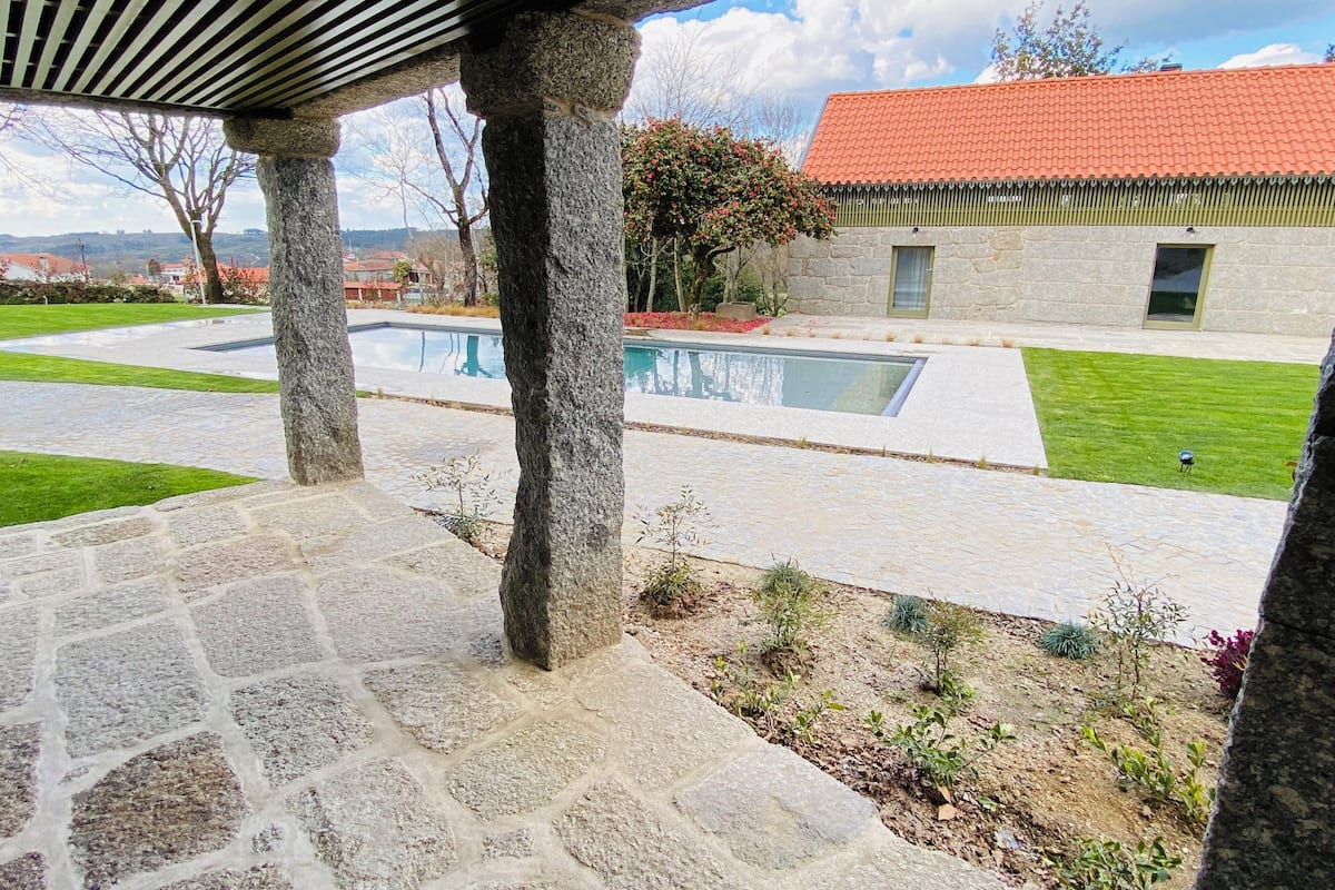 A stone-paved patio with a pool, stone pillars, and a view of a house with a red roof.