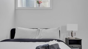A neatly made bed with white and dark grey linens, a small bedside table with a lamp, and a window with blinds and a vase of flowers. - Luxury 2Bdr Retreat, Calgary,  Everything You Need, Best coffee (Calgary)