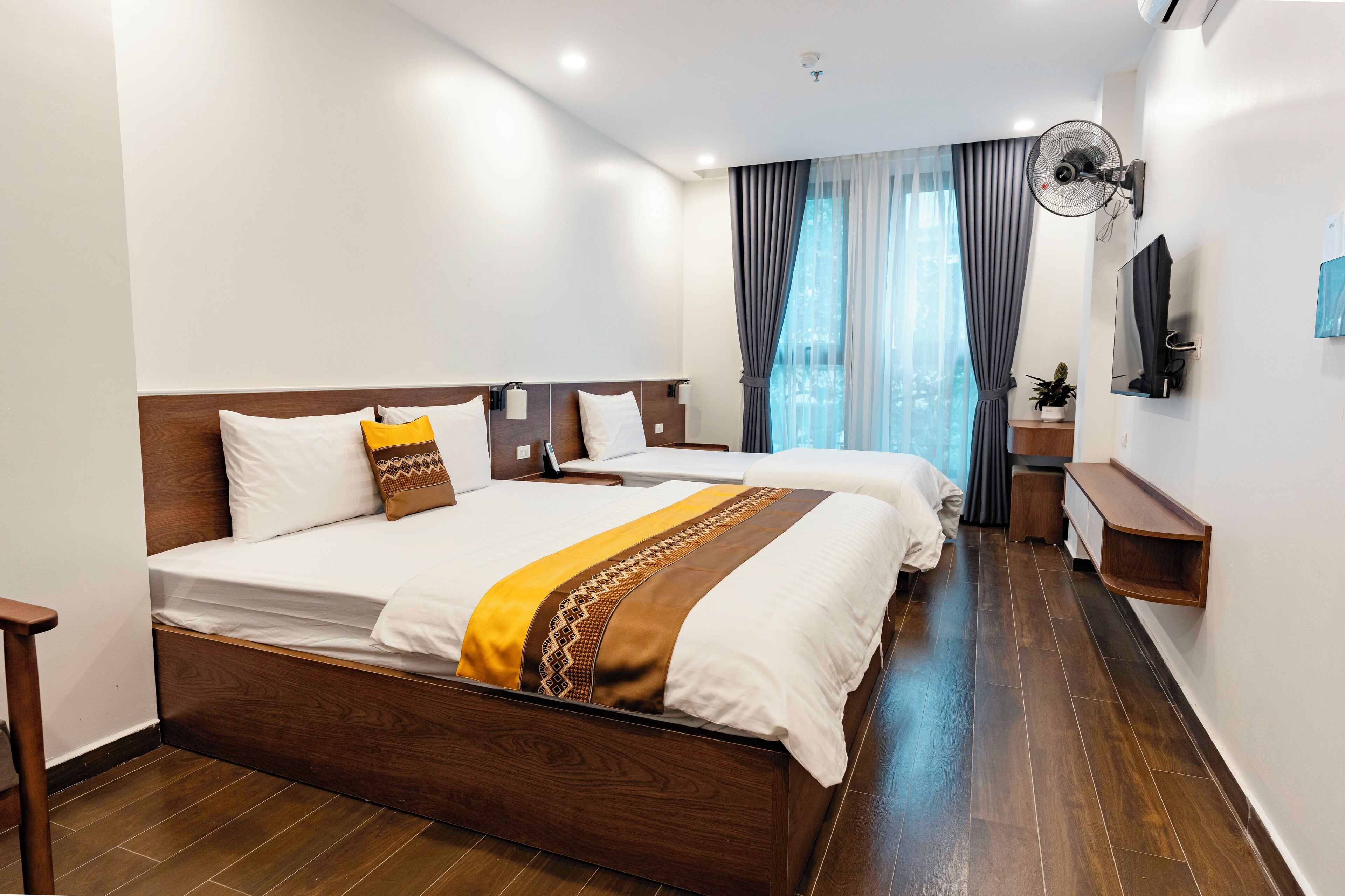 Habitación de hotel con dos camas, suelo de madera, televisor montado en la pared y ventana con cortinas.