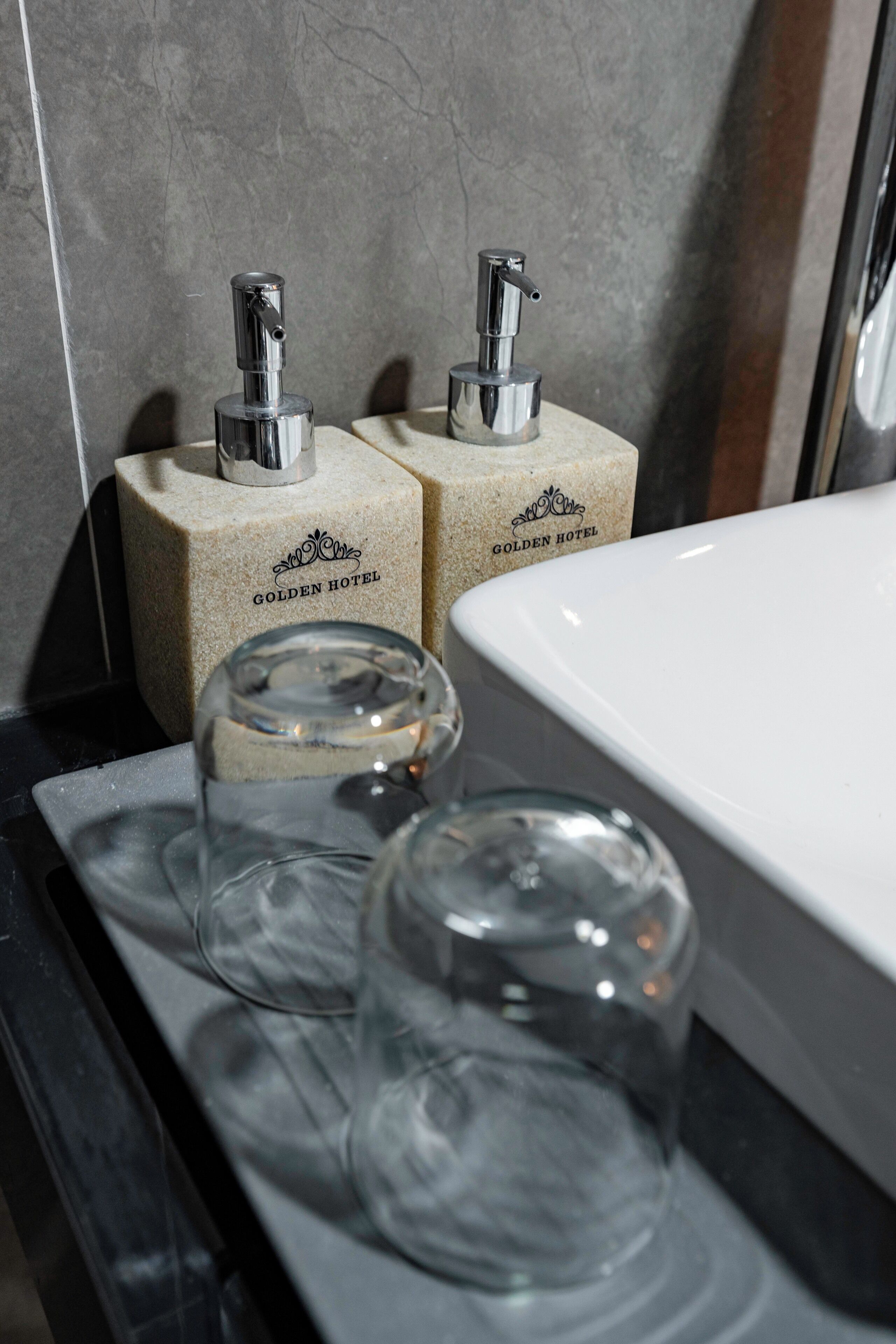 A bathroom with a white sink, two soap dispensers labeled "Golden Hotel," and two clear glass tumblers on a tray.