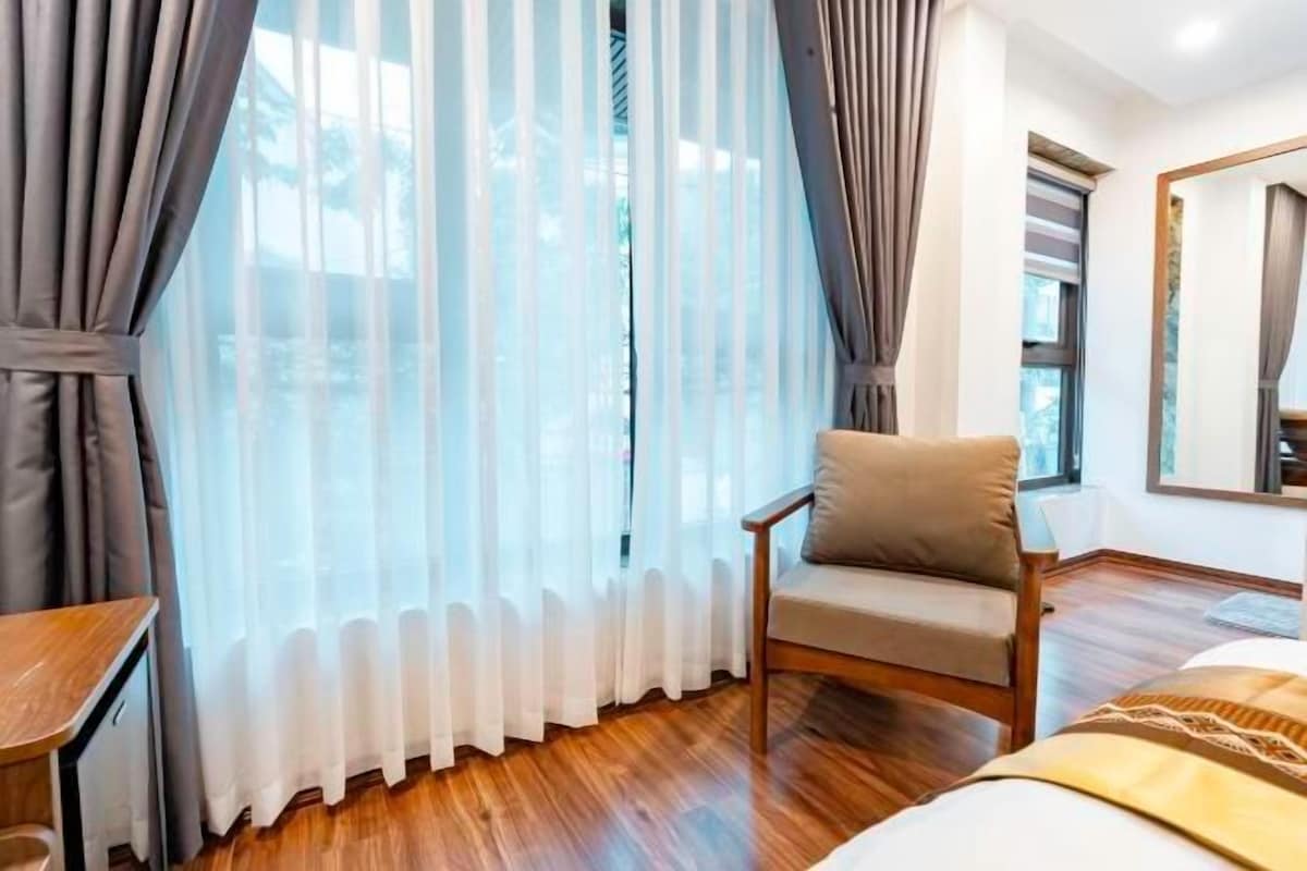 A hotel room with a wooden floor, a bed, a chair, and a window with sheer curtains.