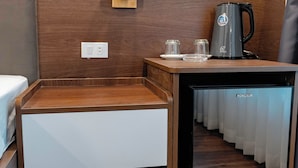 Quarto compacto com uma mesa de cabeceira de madeira, um refrigerador pequeno, uma cafeteira e uma luminária fixada na parede.