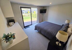 Une chambre avec un lit, un téléviseur fixé au mur, un bureau avec une lampe et une porte-fenêtre coulissante donnant sur un balcon.