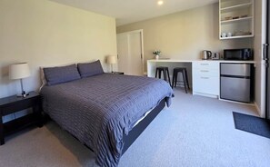 A bedroom with a bed, bedside table, lamp, and a kitchenette area with a microwave, kettle, and cabinets. - Loop the Loop (Queenstown)