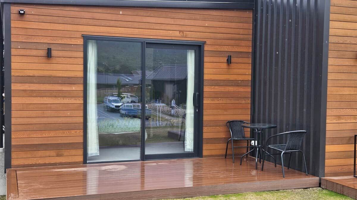 Un bâtiment moderne avec une façade en bois, une entrée dotée d’une porte en verre et un patio aménagé avec des sièges extérieurs.