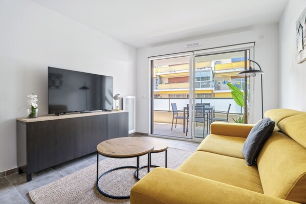 A modern living room with a sofa, a TV, a small table, and a sliding glass door leading to an outdoor balcony. - Appartement Amandiers - Welkeys (Cagnes-sur-Mer)