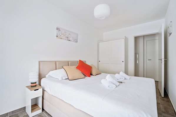 Un lit bien fait, avec une literie blanche et beige, un oreiller rouge et une table de chevet blanche sur laquelle se trouve une lampe.