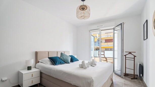 A modern bedroom with a large bed, a bedside table, a hanging light fixture, and a view of the outside balcony. - Appartement Amandiers - Welkeys (Cagnes-sur-Mer)