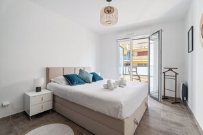 A modern bedroom with a large bed, a bedside table, a hanging light fixture, and a view of the outside balcony. - Appartement Amandiers - Welkeys (Cagnes-sur-Mer)