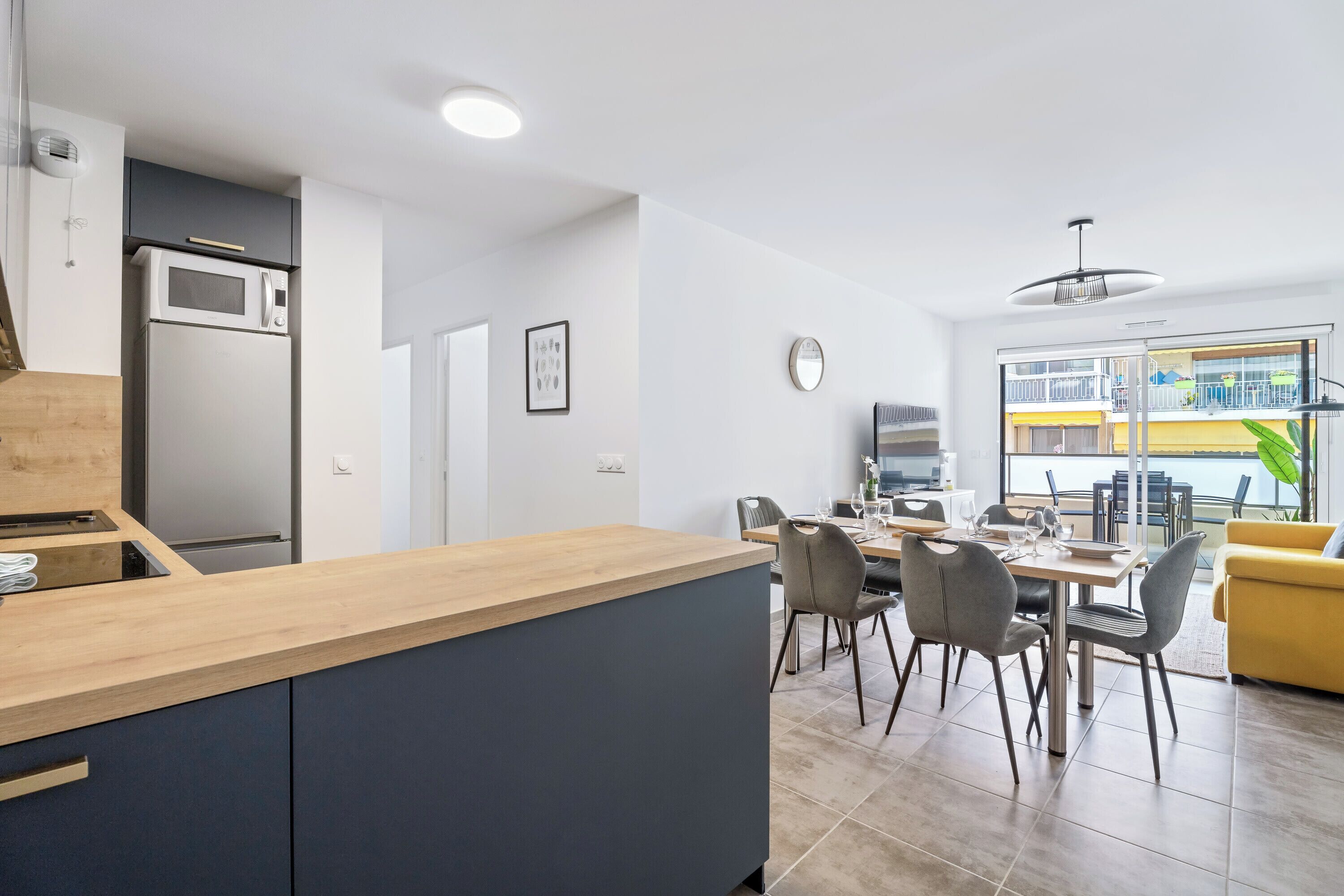 Une cuisine moderne avec un plan de travail en bois, un four à micro-ondes encastré et un réfrigérateur. On y trouve également un coin repas avec une table et des chaises, ainsi qu’un canapé jaune.
