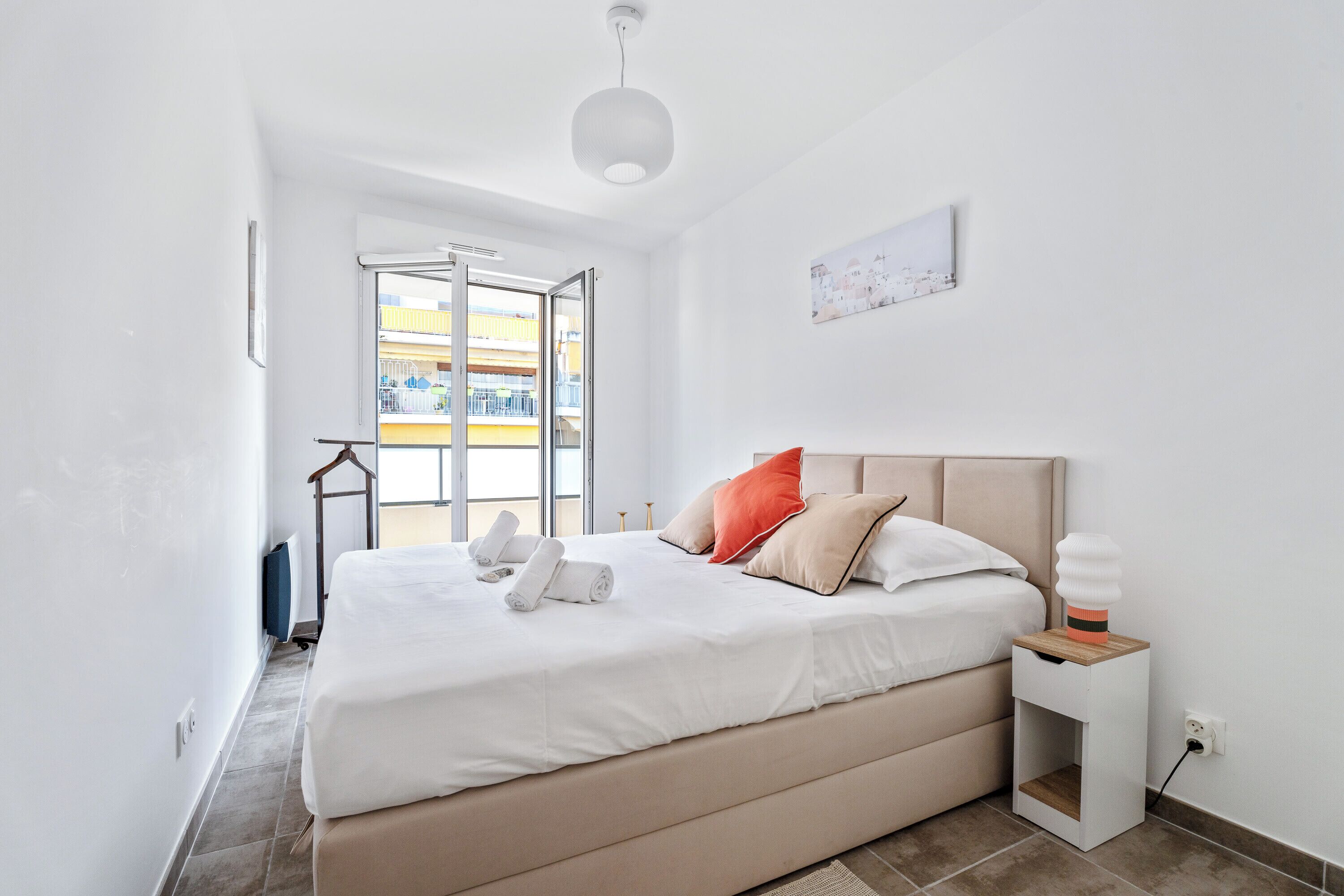 A modern bedroom with a large bed, a bedside table, a hanging light, and a view of the outside balcony.