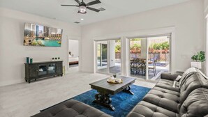 A spacious living room with a large sofa, a coffee table, and a TV mounted on the wall. There is a ceiling fan and a sliding glass door leading to an outdoor patio. - Welcome to Casa Rio, our spacious house in scenic Jensen Beach! (Jensen Beach)