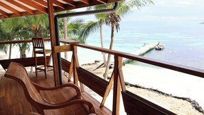 A wooden deck with two chairs overlooking a beach with palm trees and a boat docked near the shore.