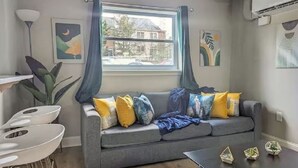 Un salon moderne avec un canapé gris, des coussins jaunes et bleus, une table blanche et une fenêtre ornée de rideaux bleus.