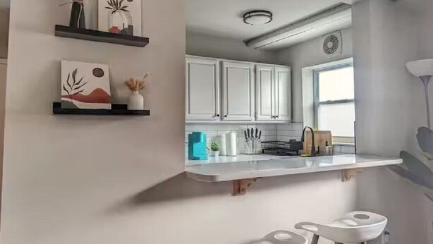 Une cuisine moderne avec des armoires blanches, un plan de travail blanc et une étagère suspendue agrémentée d’objets décoratifs.
