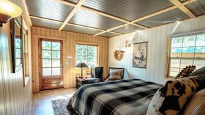 A cozy room with a wooden door, a bed with a plaid bedspread, a small table with a lamp, and a wreath on the wall. - Cattle Kate At Mountain River Ranch (Ririe)