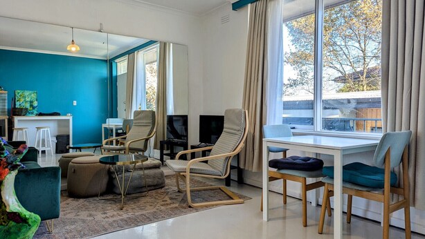 A modern living room with a blue accent wall, a dining area with a white table, and a large window with curtains. - Artist’s Studio in Fitzroy North (Fitzroy North)