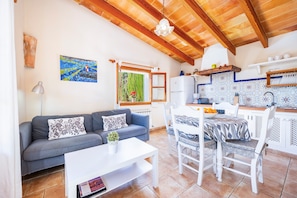 A cozy living area with a sofa, coffee table, and dining table. A kitchen with a tiled backsplash and wooden beams is visible.